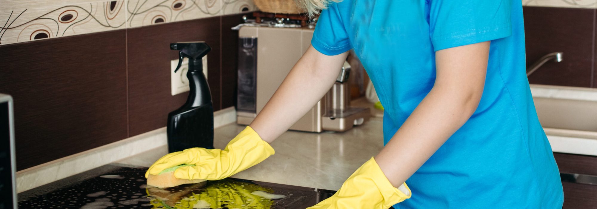Crop of blonde woman in protective gloves with rag cleaning electric stove at home kitchen. Girl washing black shiny surface of kitchen top, hand with foam on sponge. Concept of housework.